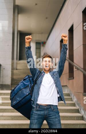 In voller Länge vertikale Porträt von fröhlich gut aussehenden jungen Mann der Lebensmittelzustellung Service mit großen thermischen Rucksack heben die Hände, den Sieg zu feiern Stockfoto