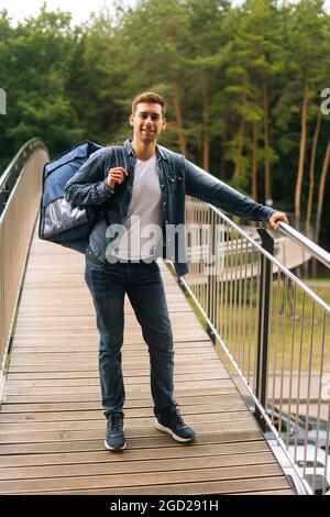 In voller Länge vertikale Porträt von fröhlich hübsch jungen Mann der Lebensmittelzustellung Service mit großen thermischen Rucksack auf der Terrasse des Balkons stehen. Stockfoto