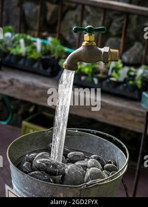 Wasserhahn im Garten mit magischer Erhaltung: Wasserlauf mit neuem Trick, Wasser fließt in Stein gefüllte Wasserkübel aus Wasserhahn, der in der Luft schwimmt Stockfoto