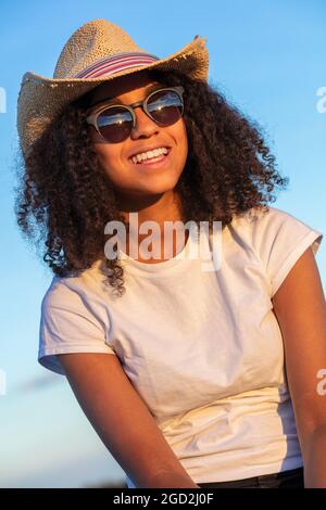 Schöne und glückliche gemischte Rasse afroamerikanische weibliche Mädchen Teenager junge Frau mit perfekten Zähnen tragen Stroh Cowboy Hut Sonnenbrille und weißen t-sh Stockfoto