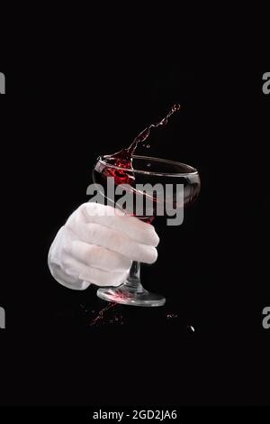 Glas Rotwein mit fliegenden Spritzern mit der Hand eines Kellners in einem weißen Handschuh auf schwarzem Hintergrund, verschüttete Getränke, Tropfen. Stockfoto