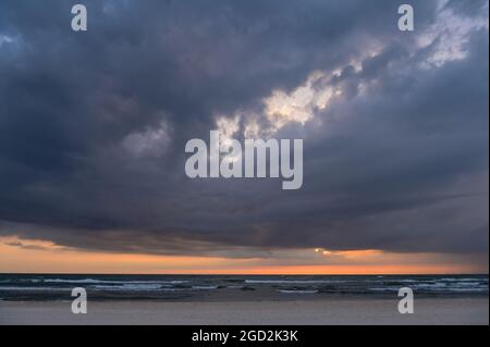 Dunkle Wolken bei Sonnenuntergang am Meer mit kleinen Wellen und die Sonne verstreut hinter den Wolken nur einen orangen Streifen am Horizont Stockfoto