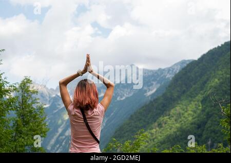 Blick von hinten auf eine junge Frau, die in wunderschöner Natur steht, mit ihren Händen, die in einer betenden Position über ihrem Kopf zusammengeschlossen sind. Stockfoto