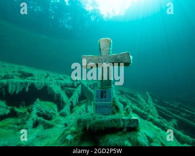 Steinkreuz unter Wasser in einem Bergsee, gute Sicht, klares Wasser und einige Baumstämme unter Wasser, Spiegelung eines Waldes auf der Wasseroberfläche, Stockfoto