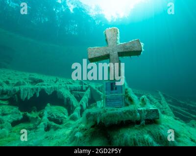 Steinkreuz unter Wasser in einem Bergsee, gute Sicht, klares Wasser und einige Baumstämme unter Wasser, Spiegelung eines Waldes auf der Wasseroberfläche, Stockfoto