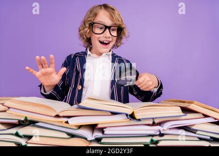 Portrait von trendy fröhlich Schuljunge lesen Haufen Buch Lösung mit Schleife isoliert über lila violetten Hintergrund zu finden Stockfoto