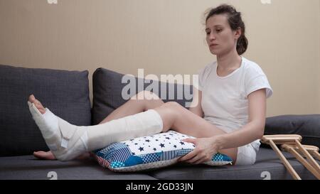 Eine Frau mit Gipsabdruck sitzt auf einem Sofa, ein Kissen wird unter ein gebrochenes Bein gelegt. Stockfoto