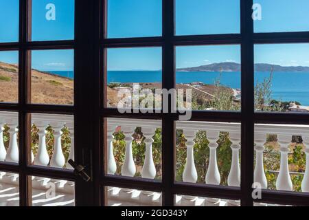 Traditionelle griechische Architektur, weißer Balkon mit Balustern. Blick von der geschlossenen Glastür auf die klare blaue Küste der Ägäis. Sommer landschaftlich schöne Aussicht Fr. Stockfoto