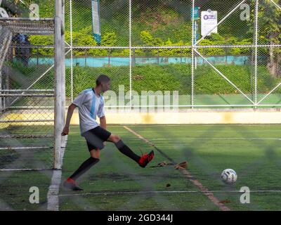 Medellin, Antioquia, Kolumbien - Juli 30 2021: Der lateinische Mann in Sportswear läuft auf einem grünen Synthetikfeld, um einen Ball in der Nähe eines Fußballtals zu schießen Stockfoto