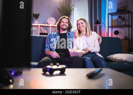 Positives junges Paar, das sich im Fernsehen einen Film ansieht und zu Hause Popcorn isst. Glücklicher kaukasischer Mann und Frau, die auf der Couch sitzen und umarmen. Zeit zusammen. Stockfoto
