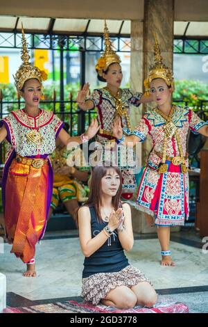 Attraktive Thai-Frau, die im Erawan-Schrein betet, mit prunkvollen Thai-Tänzern im Hintergrund, Bangkok, Thailand Stockfoto