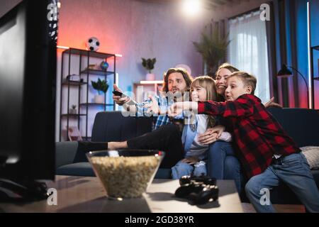 Fröhliche Eltern mit zwei Kindern, die in Umarmungen auf der gemütlichen Couch sitzen und die Fernbedienung benutzen, während sie Fernsehen. Glückliche Familie mit echten Emotionen auf Gesichtern zu Hause. Stockfoto