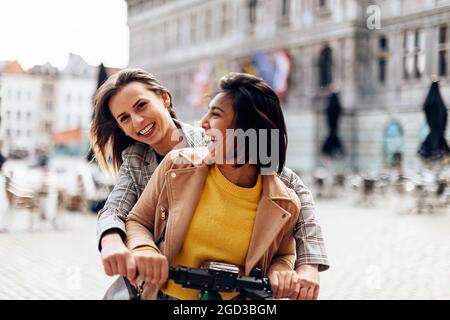 Lächelnde multirassische Freundinnen mit Spaß auf einem Elektroroller. Stockfoto