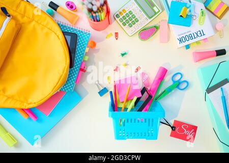 Zurück zur Schule. Obere Ansicht des weißen Schreibtisches mit Arbeitsbüchern, Schreibwaren, Lehrbuch, Rucksack, Verbraucherkorb und Verkaufsetikett im Schulkinderzimmer in der Sonne Stockfoto