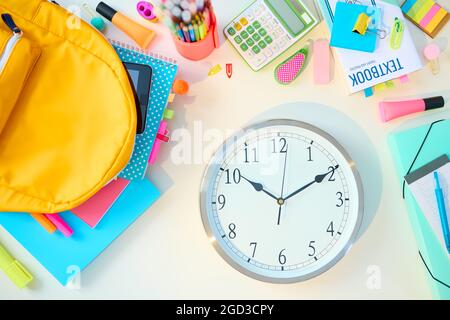 Zurück zur Schule. Obere Ansicht des weißen Tisches mit Arbeitsbüchern, Schreibwaren, Lehrbuch, Uhr und Rucksack im Studentenzimmer an sonnigen Tagen. Stockfoto