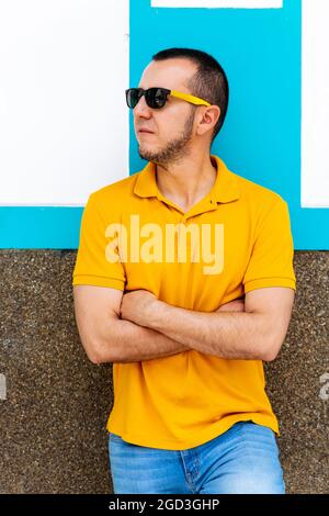 Vertikale Aufnahme eines jungen Latino-Mannes mit Sonnenbrille und gekreuzten Armen an einer blau-weißen Wand. Stockfoto
