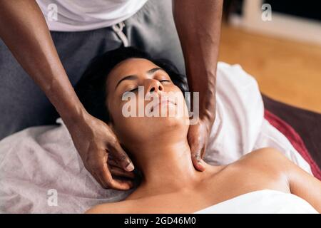 Entspannte Frau, die sich hinlegt und eine Nackenmassage erhält. Die Masseuse ist ein hübscher afroamerikanischer Mann. Stockfoto