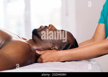 Entspannter schwarzer Mann, der sich hinlegt und eine Nackenmassage erhält. Die Masseuse verwendet eine Ölmassage. Stockfoto