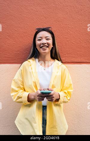 Junge und hübsche asiatische Frau, die lächelt und die Kamera anschaut, während sie ihr Mobiltelefon hält. Stockfoto