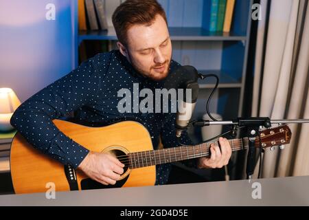 Mittellange Aufnahme eines gespielten Gitarristen-Sängers, der auf einer akustischen Gitarre spielt und in ein Mikrofon singt, um im Heimstudio einen Song aufzunehmen.