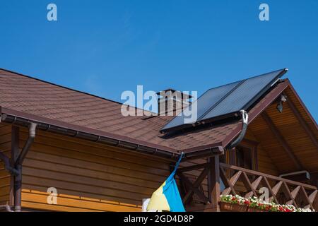 Solarthermische Einzelheizung für Warmwasser auf Bitumen-Asphalt-Schindeln Dach. Gemauerter Schornsteinrohr auf einem Holzhaus. Regenrinne. Stockfoto