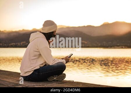 Nicht erkennbare junge Frau in weißem Wollhut sitzt auf einem Pier allein tragen Schutzmaske mit Smartphone in Kontakt mit Freunden und f zu halten Stockfoto