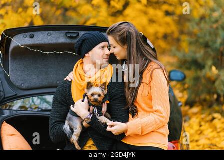 Ein junges verliebtes Paar sitzt mit seinem kleinen Hund auf dem offenen Kofferraum eines schwarzen Autos im Herbstwald. Liebhaber schauen sich gegenseitig an Stockfoto