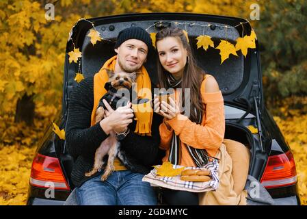 Ein junges Paar, ein Mann und eine Frau, in warmen Kleidern gekleidet, sitzen auf dem Kofferraum eines Autos und trinken heißen Tee an einem hellen Herbsttag im Wald mit Stockfoto