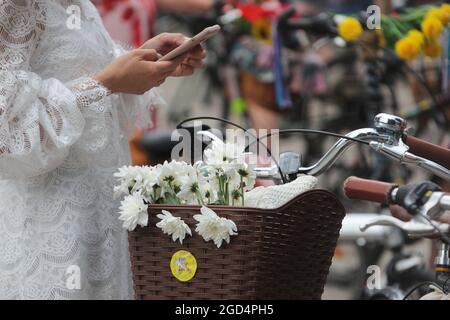 KIEW, UKRAINE - 7. AUGUST 2021 - EINE Frau in einem weißen Spitzenkleid stöbert ihr Smartphone neben einem Korb mit weißen Blumen, die an einem Fahrrad befestigt sind, siehe Stockfoto