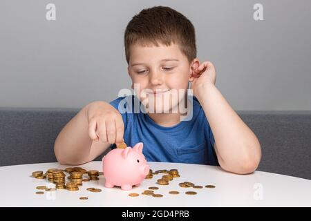 Das Kind schaut auf das Sparschwein und legt Münzen ab. Der Junge lächelt, während er investiert. Stockfoto