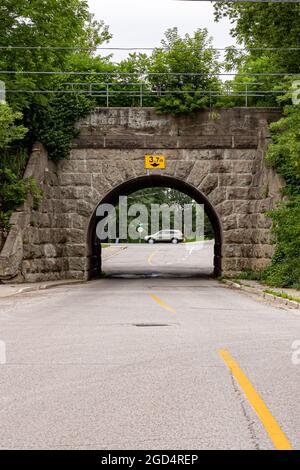 Vertikaler selektiver Fokus auf Steinautobahn unter einer Eisenbahnbrücke. Unscharfes Auto im Hintergrund, Straßenlinie im Vordergrund. Stockfoto