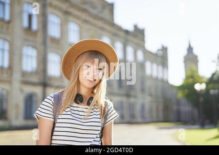 Fröhlich lächelnd junge blonde europäische Universitäts- oder Hochschulstudentin mit braunem Hut auf dem Campus. Nettes Schulmädchen, Studentenportrait oder Bildungskonzept. Hochwertige Bilder Stockfoto