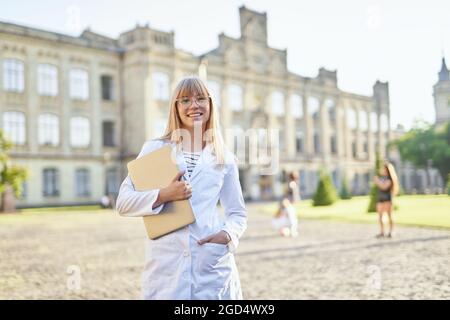 Porträt eines jungen blonden Medizinstudenten mit Laptop, der im Freien in einem weißen medizinischen Kleid steht. Praktikanten oder studentische Ärztin auf dem Universitätscampus schauen auf die Kamera. Bildungskonzept Stockfoto