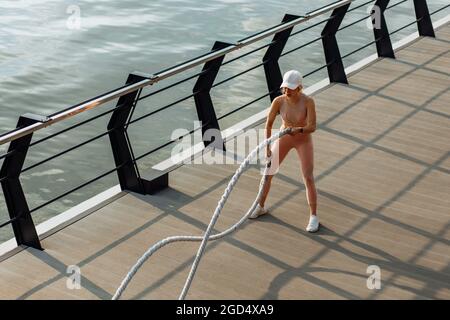 Fitness-Frau mit Trainingsseilen für den Outdoor-Sport am Pier, Sportswoman-Training mit Kampfseilen, Sportkonzept, gesunder Lebensstil Stockfoto