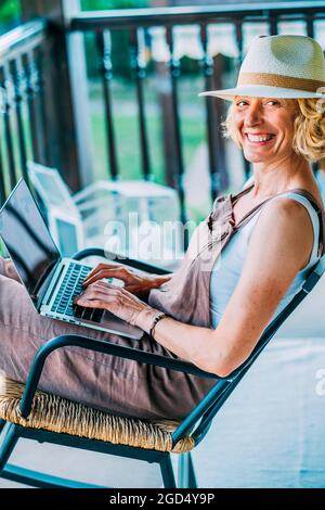 Reife kaukasische blonde Frau in den 50er Jahren mit einem Laptop mit Hut und sitzt in einer entspannten Pose in einem Schaukelstuhl auf der Veranda. Lifestyle-Konzept. Stockfoto