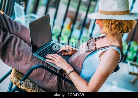 Reife kaukasische blonde Frau in den 50er Jahren mit einem Laptop mit Hut und sitzt in einer entspannten Pose in einem Schaukelstuhl auf der Veranda. Lifestyle-Konzept. Stockfoto