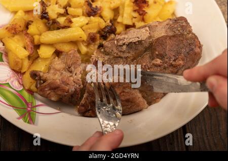 Jemand schneidet mit einem Messer und einer Gabel ein Stück saftiges Braten auf einem Teller gebratener Kartoffeln. Nahaufnahme Selektiver Fokus Stockfoto