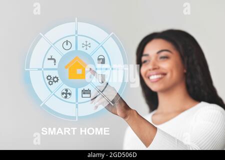 Glückliche Millennial schwarze Dame berühren runde virtuelle Schnittstelle Smart Home-Bildschirm und lächelnd, genießen moderne Technologien, Steuerung Elektronik, Stockfoto