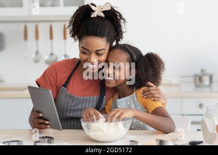 Lächelnde afroamerikanische Mutter und Kind Teenager-Mädchen halten digitale Pad beim Backen in der Küche zusammen, Selfie auf Pad beim Kochen oder mit V Stockfoto