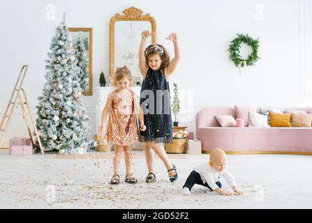 Zwei Mädchen und ein kleiner Junge spielen mit Weihnachtskonfetti im Wohnzimmer neben dem Weihnachtsbaum Stockfoto