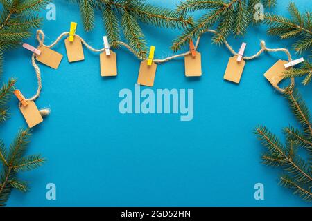 Papier-Tags mit farbigen Wäscheklammern an Jute Seil gebunden, Seil mit Tags auf blauem Hintergrund mit Tannenzweigen, weihnachten oder Neujahr Hintergrund Stockfoto