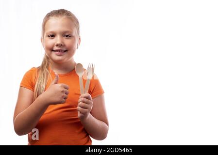 Niedliches kaukasisches kleines Mädchen, das Bambusbesteck in den Händen hält und Daumen auf weißem Hintergrund zeigt. Bambus-Holzbesteck. Zero Waste Konzept, Stockfoto