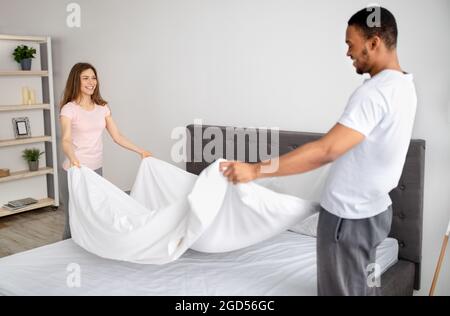 Fröhliches, junges multinationales Paar, das zu Hause Bett macht, eine Decke hält und gemeinsam Hausarbeit leistet. Liebevolle Millennial-Familie putzt ihr Schlafzimmer, Stockfoto