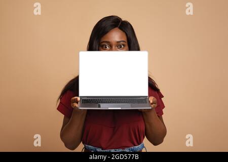 Junge schwarze Dame hält Notizbuch mit leerem Bildschirm, beigem Studiohintergrund, Mockup, Kopierraum. afroamerikanische Frau versteckt sich hinter dem Computer mit b Stockfoto