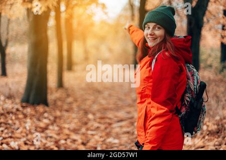 Weibliche Reisende mit Rucksack zeigt die Fahrtrichtung, Blick von hinten, Wandern im Wald, Herbstlandschaft Stockfoto
