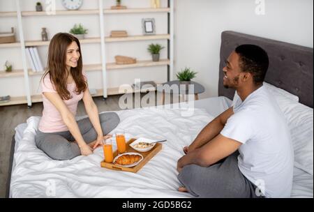 Der junge schwarze Kerl und seine Freundin frühstücken zu Hause auf dem Bett. Liebevolles interrassisches Paar, das zusammen am faulen Morgen leckeres Essen isst, enjoyi Stockfoto