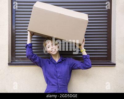 Eine junge lächelnde Frau in lila Uniform hält einen Karton auf ihrem Kopf. Lieferung, Überraschung, schöner Kurier. Geschäft, Post, Geschenk. Teilzeitjobs Stockfoto