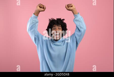 Triumphender schwarzer Teenager, der die Arme über seinen Kopf hebt und vor Aufregung auf rosa Hintergrund schreit. Entzückende afroamerikanische Jugendliche feiern su Stockfoto