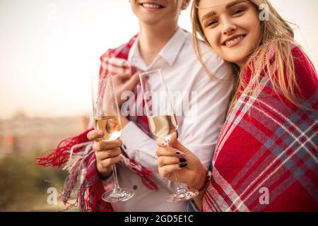 Ein wunderschönes, liebevolles Paar, in eine Decke gehüllt, lächelnd auf die Kamera, ein junger Mann und eine junge Frau halten Champagner in der Hand, während sie am freitag gemeinsam Zeit genießen Stockfoto