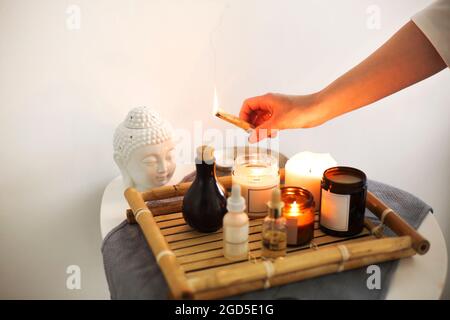 Weibliche Hand, die Räucherstäbchen über einem Korbtablett mit Kerzen, verschiedenen Ölen und Cremes auf DEM SPA-Tisch hält, der mit einem kleinen Buddha-Kopf in Massage dekoriert ist Stockfoto